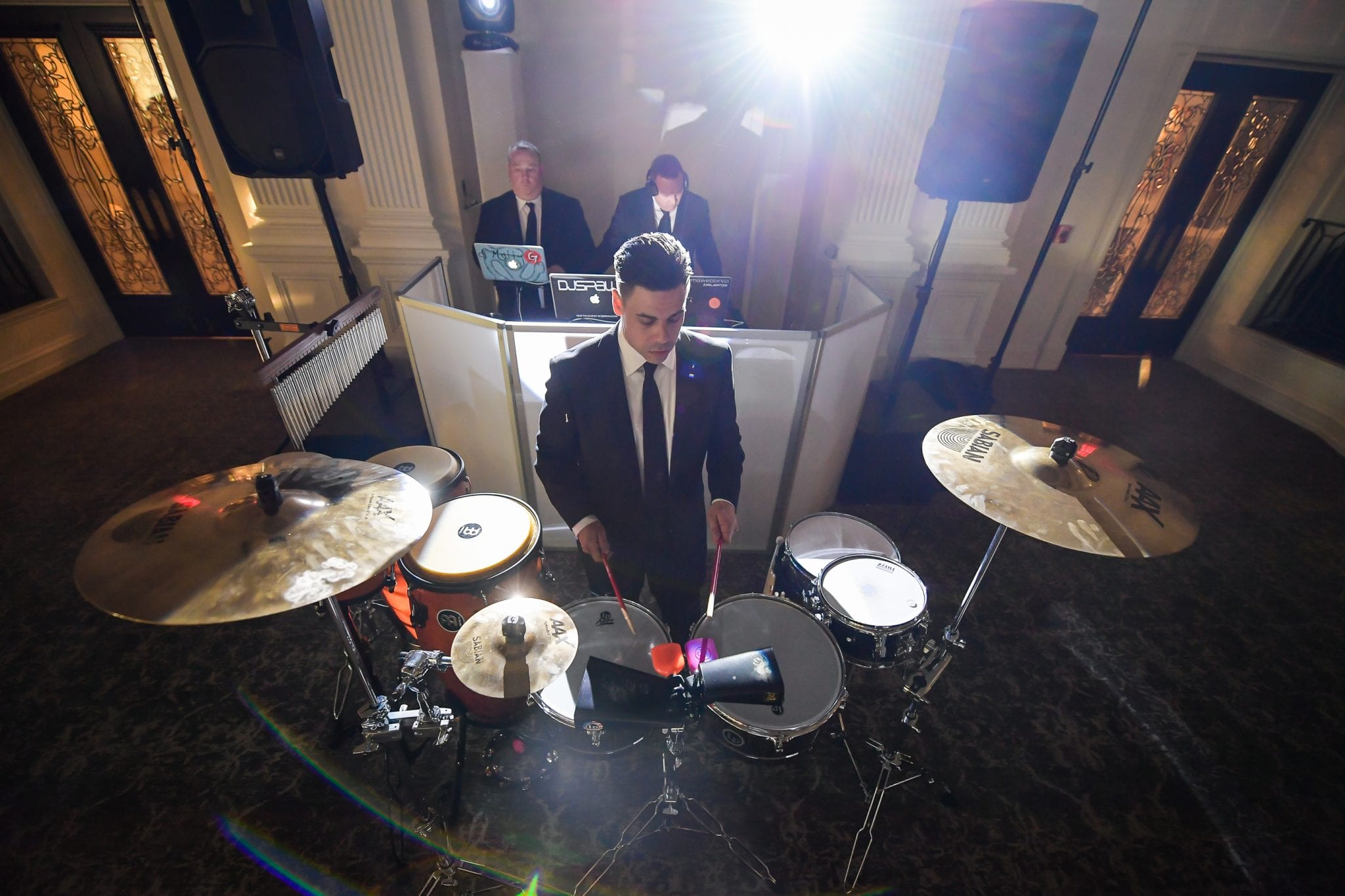 A drummer playing in front of a set of DJs in a DJ booth behind him.